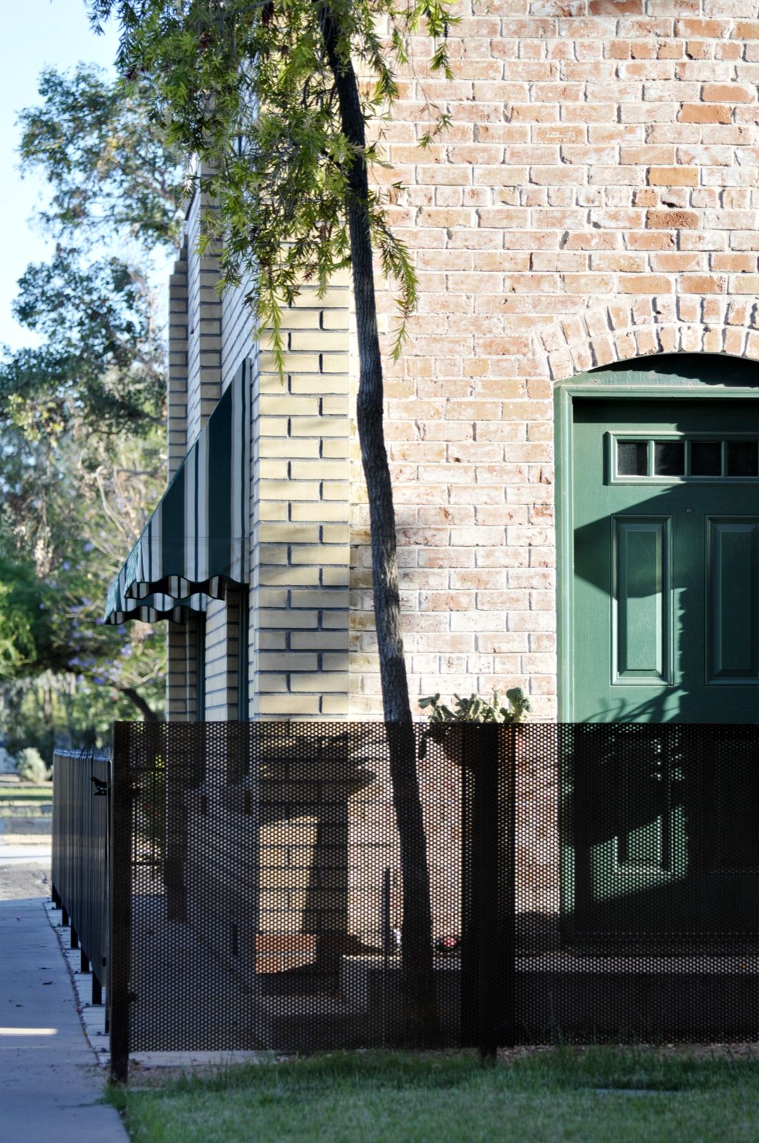Fence detail of perforated steel sheet in the foreground and historic brick wall house with green-white shade canopies in the background